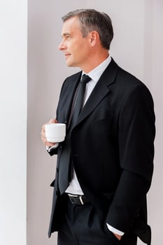 Relaxing during coffee break. Thoughtful mature man in formalwear holding coffee cup and looking away while standing near window photo