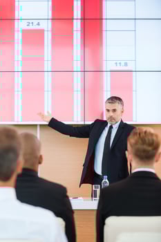 Talking about company achievements. Confident mature man in formalwear pointing projection screen with graph on it while making presentation in conference hall with people on the foreground photo