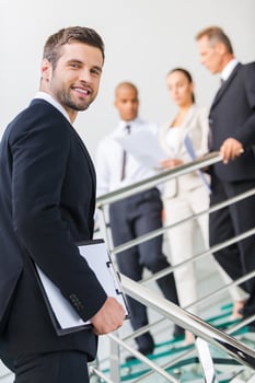 Young and successful. Low angle view of confident young man in formalwear looking over shoulder and smiling while moving up by staircase with people in the background photo