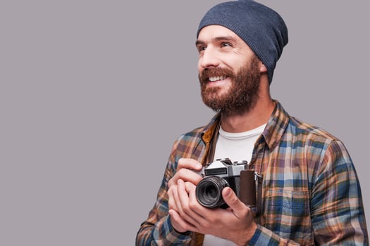 Happy photographer. Handsome young bearded man holding old-fashioned camera and looking away with smile while standing against grey background photo