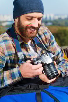 Remembering the whole trip.  Handsome young man sitting near backpack and holding camera photo