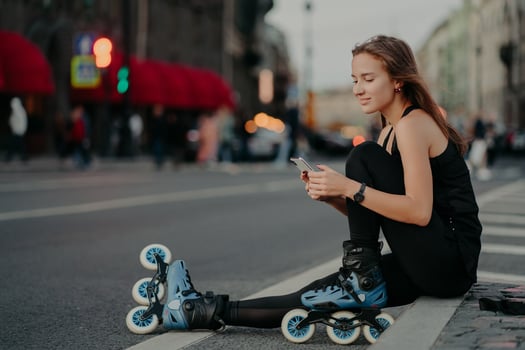 Outdoor shot of sporty active young woman dressed in active wear wears rollerblades checks message content on smartphone poses against blurred city background on asphalt leads healthy lifestyle photo