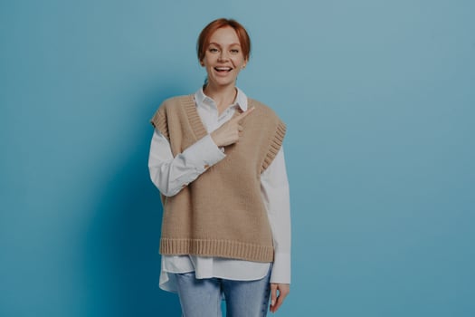 Positive stylish red-haired woman in stylish casual outfit pointing at copy space, isolated on blue photo