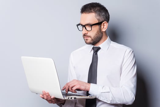 Business expert at work. Confident mature man in shirt and tie working on laptop while standing against grey background photo