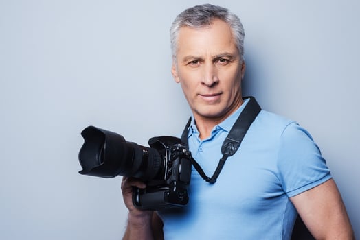 Professional photographer. Portrait of confident mature man in T-shirt holding camera while standing against grey background photo