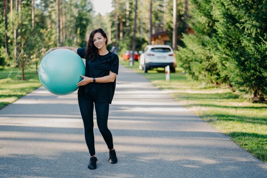 Full length shot of active sporty woman holds big fitness ball exercises outdoor walks on road near forest leads healthy lifestyle stays fit dressed in sportswear. People gymnastics and aerobics photo
