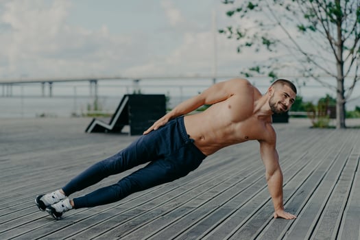 Sporty bodybuilder stands in side plank, exercises outdoor and wears sport clothes, leads healthy lifestyle, has fit body and muscles. Outdoor shot of determined sportsman does health activities photo