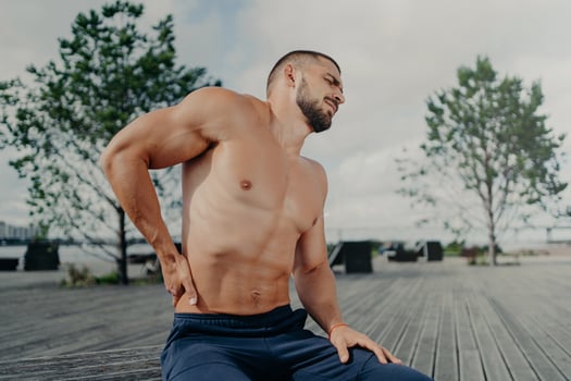 Displeased unshaven European sportsman touches lower back and feels backache after physical training exercises, poses outdoor. Sport exercising injury. Athletic guy suffers pain, needs massage photo