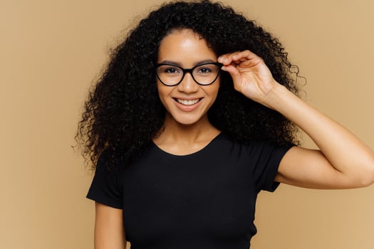Lovely young African American woman grins at camera, keeps hand on rim of spectacles, glad to hear good news, dressed in casual black t shirt, isolated over brown background. Happiness concept photo