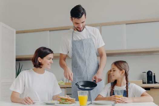 Busy caring father prepares delicious breakfast for wife and daughter, wears apron, puts fried eggs on plate. Cheerful mother and child talk to each other while sit at kitchen table, have meal photo