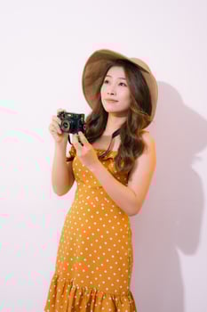 Pretty young tourist woman in summer white dress hat with photo camera isolated on pink background.