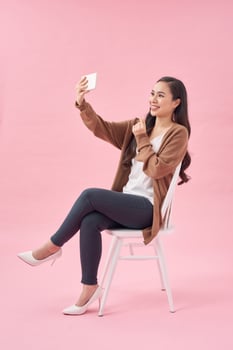 Smiling young woman making selfie photo on smartphone over pink background