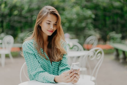Beautiful woman with makeup, dressed in fashionable clothes, dressed in fashionable polkadot shirt, uses mobile phone for online communication, surfes social networks, poses in outdoor cafeteria photo