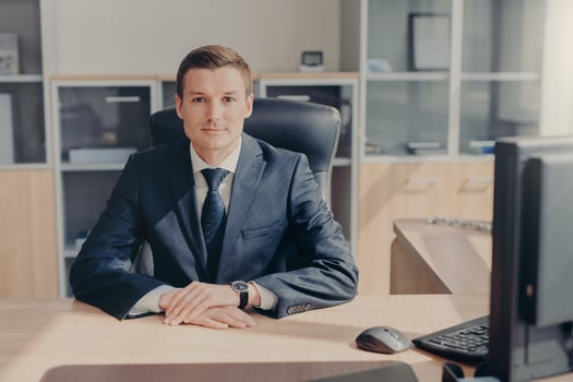 Horizontal shot of prosperous young businessman in elegant suit, sits at work place, surrounded with computer, has confident look directly into camera, being professional in business sphere. photo