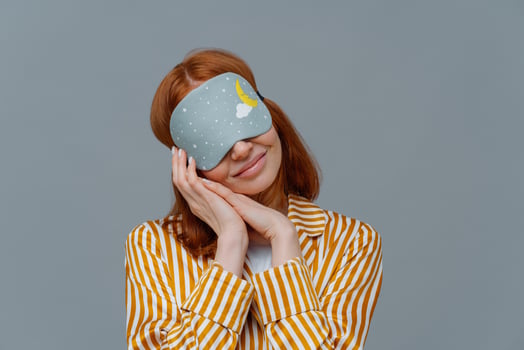 Pretty redhead woman wears blindfold on eyes, keeps hands pressed together near face, sees pleasant dreams while sleeping, dressed in domestic clothes, isolated on grey wall, feels comfortable photo