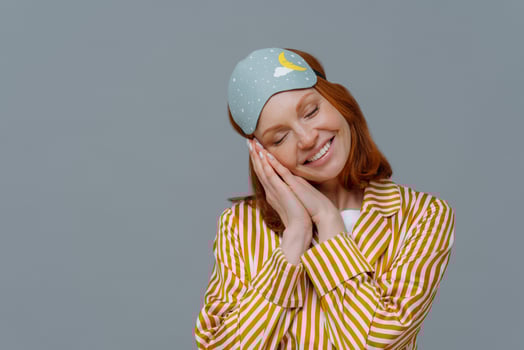 Deep sleep and pleasure. Pretty freckled woman tilts head, smiles broadly, shows white teeth, wears domestic clothes and blindfold, sees pleasant dreams, has nice day, doesnt want to wake up photo