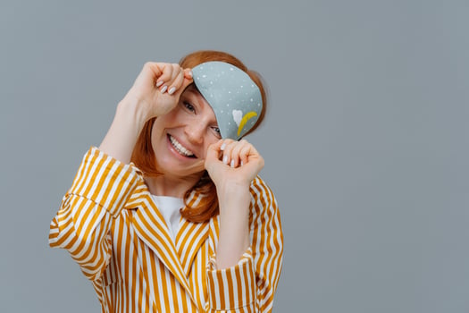 Positive good looking woman with toothy smile, has nice dream, smiles pleasantly, wears blindfold and pajamas, awakes after seeing good dreams, stands over grey studio wall with blank copy space photo