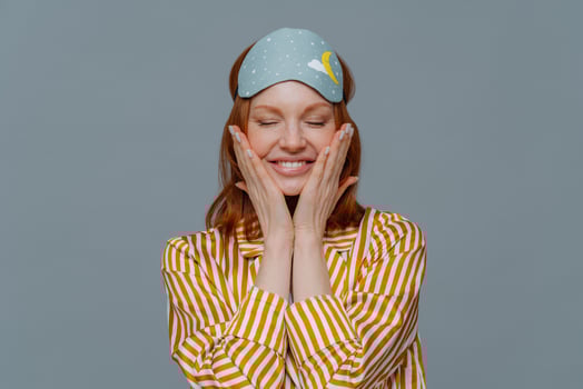 Pleased woman touches cheeks, has healthy freckled skin, smiles broadly prepares for sleep, wears striped pajama and sleep mask, closes eyes, isolated over grey background. Rest and lifestyle concept photo