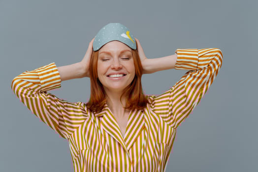 Daytime nap concept. Pleased woman with ginger hair wears comfortable striped pajama and sleep mask, uses useful accessory for sleep, smiles broadly, shows white teeth, isolated on grey studio wall photo