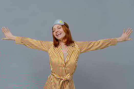 Positive relaxed woman wears blindfold and striped robe stretches arms sideways, smiles broadly, closes eyes, isolated over grey background, wakes up in bedroom, slept well. Good morning concept photo