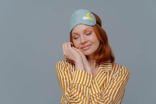 Pretty foxy freckled woman leans at palms, keeps eyes closed, wears sleepmask and stiped pajamas, dreams about something pleasant, stands indoor over grey wall, with copy space for promotion photo