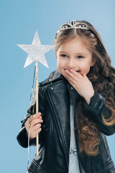 Glad pleasant looking small kid covers mouth with palm, giggles positively, wears crown and black leather jacket, holds magic wand, isolated over blue background. Children and happiness concept photo