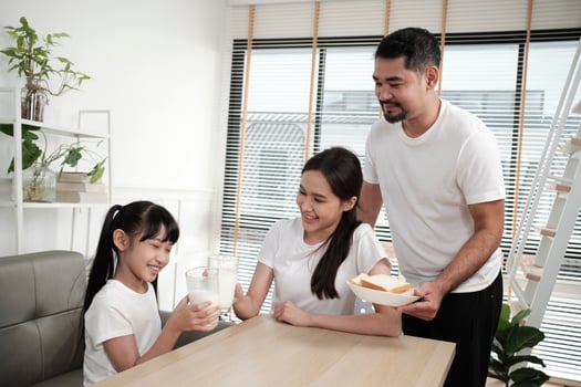 A Healthy Asian Thai family, daughter, and young mum drink fresh white milk in glass and bread from father, joy together at a dining table in morning, wellness nutrition home breakfast meal lifestyle. photo