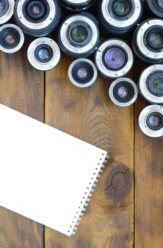 Several photographic lenses and white notebook lie on a brown wooden background. Space for text photo