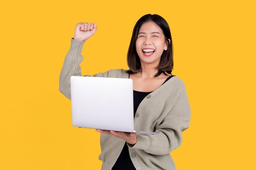 Happy smiling Asian woman wearing casual clothes is working on a laptop PC computer gesturing winner isolated on yellow background. People lifestyle concept. photo