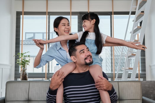 Asian Thai family together, dad plays and teases with daughter and mum by carrying and holding girl on shoulders in home living room, happy leisure times, lovely weekend, wellbeing domestic lifestyle. photo
