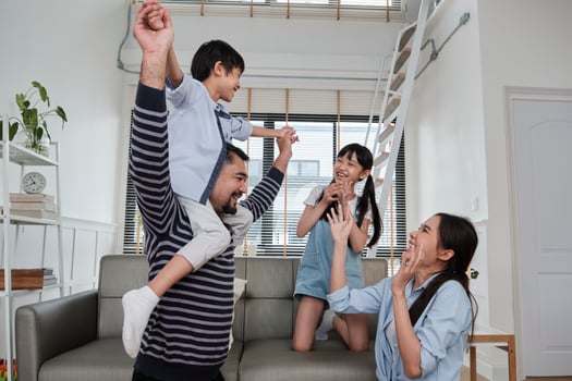 Asian Thai family together, dad plays and teases with children and mum by carrying and holding son on shoulders in home living room, happy leisure times, lovely weekend, wellbeing domestic lifestyle. photo