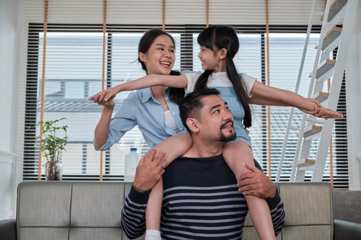Asian Thai family together, dad plays and teases with daughter and mum by carrying and holding girl on shoulders in home living room, happy leisure times, lovely weekend, wellbeing domestic lifestyle. photo