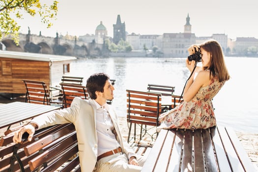 Professional beautiful female photographer makes photo of her husband who poses at camera on wooden bench at outdoor cafe near beautiful panoramic ancient city view. People and hobby concept