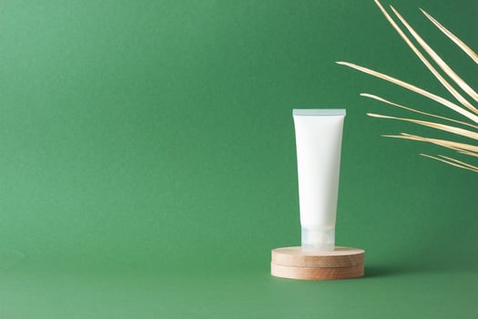 White tube of cosmetic cream with wood podium and palm leaf on green background. Close up, copy space photo