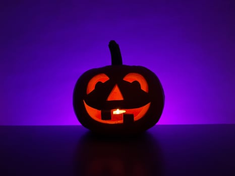 Carved glowing pumpkin jack-o-lantern with candle inside stands on surface in dark night and blurred defocused purple background. The back wall highlighted in violet. Halloween holiday concept. photo