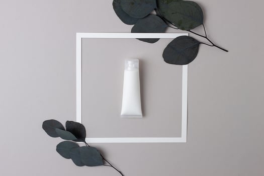 White tube of cosmetic cream with white frame and eucalyptus on grey background. Flat lay photo