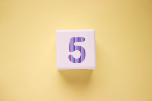 Close-up photo of a white plastic cube with number 5 on a yellow background. Object in the center of the photo
