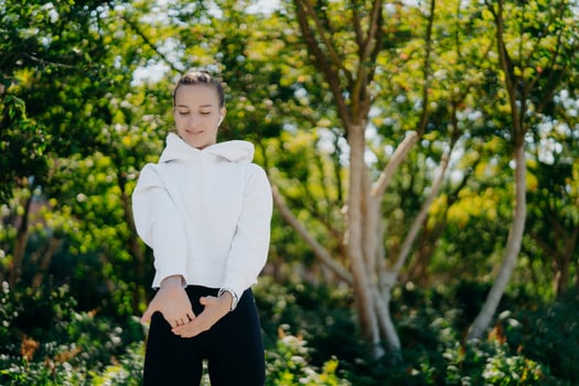 Horizontal shot of sporty European woman stretches hands and warms up before workout wears white sweatshirt andleggings poses against trees background spends free time outdoors on fresh air. photo