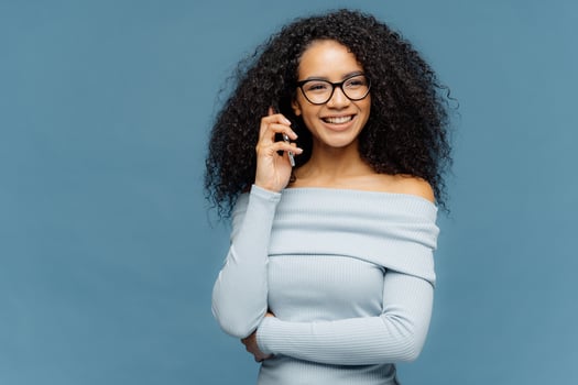 Indoor shot of positive lovely woman has telephone conversation, keeps mobile phone near ear, focused aside, wears casual jumper, isolated on blue background. People, technology, communication photo