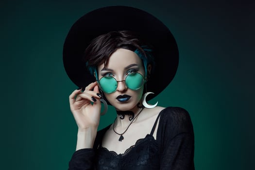 Young Halloween witch in a round hat and green round glasses on a green dark background. Dyed hair, dark lipstick, crescent moon earrings photo