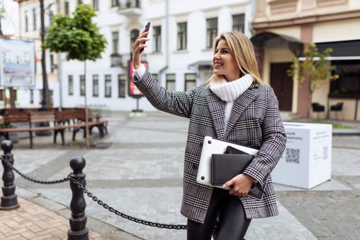 stylish business adult woman with a laptop in her hands takes a selfie on a mobile phone near the city square photo