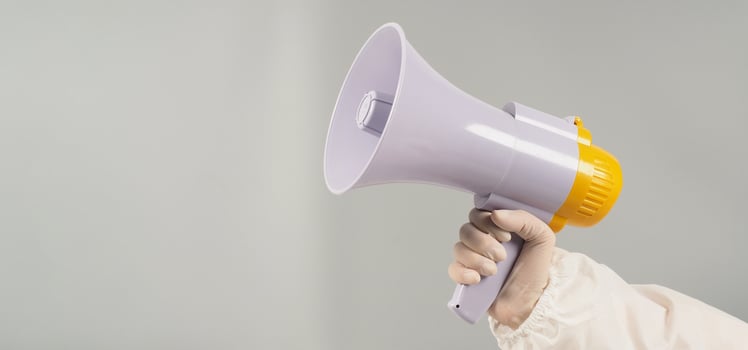Hand is holding megaphone on grey background. Hand wears a PPE suit and a white medical glove. photo