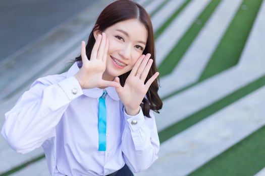 Beautiful high school Asian student girl in the school uniform stands and smiles happily with braces on her teeth while she put her hand on face confidently while she looks at the camera photo