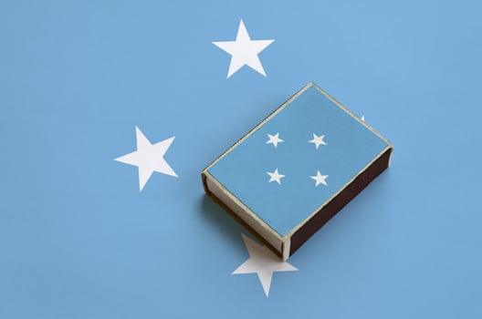 Micronesia flag is pictured on a matchbox that lies on a large flag photo