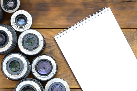 Several photographic lenses and white notebook lie on a brown wooden background. Space for text photo