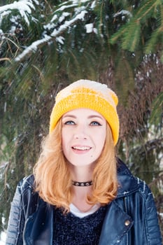 Young woman with blue eyes and blond hair in a yellow knitting hat and black leather jacket under the fir tree at the moment of snowfall. photo