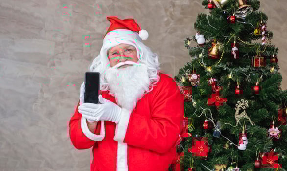 Santa claus celebrating Christmas in happiness and excitement while holding mobile phone beside the fully decorated christmas tree for sale promotion and online shopping concept photo