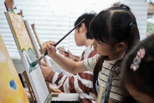 A young Asian girl concentrates on acrylic color picture painting on canvas with student kids in an art classroom, creative learning with talents and skills in the elementary school studio education. photo