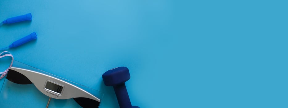 Woman measures her weight on scale on a blue background with skipping rope and dumbbells. Healthy lifestyle banner photo