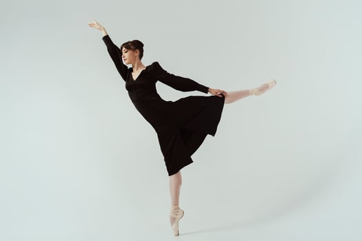 ballerina in a black dress and ballet shoes poses in a photo studio in motion showing beautiful long legs make attitude